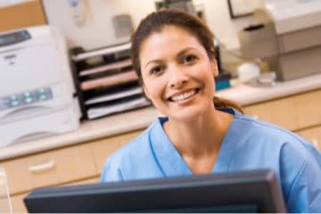 A young smiling woman in blue scrubs sitting in front of a computer