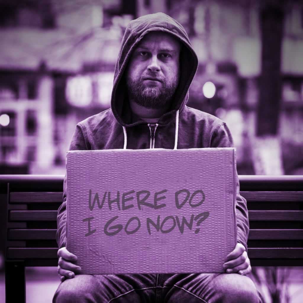 Homeless person sitting on a bench holding a sign that says "Where do I go now?"