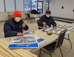 Two men sit at table as they construct picket signs. One in a red tuque holds up his thumb